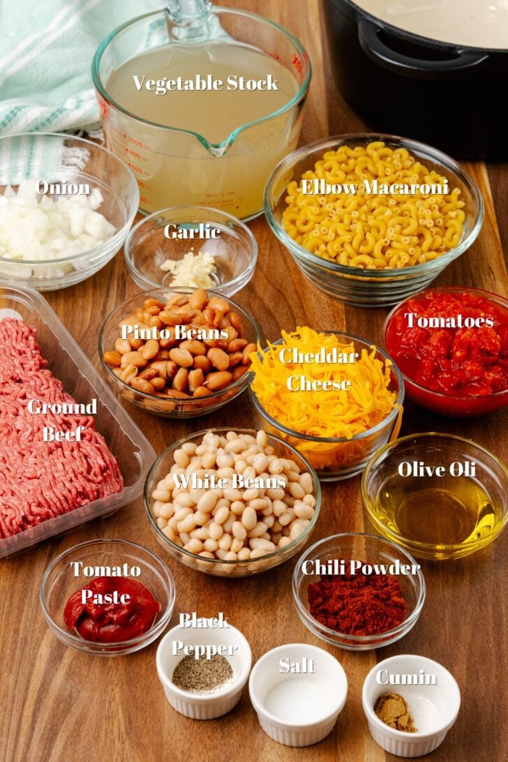 Ingredient list for cheesy chili mac including ground beef, seasonings, white beans, pinto beans, cheddar cheese, chopped tomatoes, elbow macaroni, vegetable stock, and chopped onions in glass bowls.