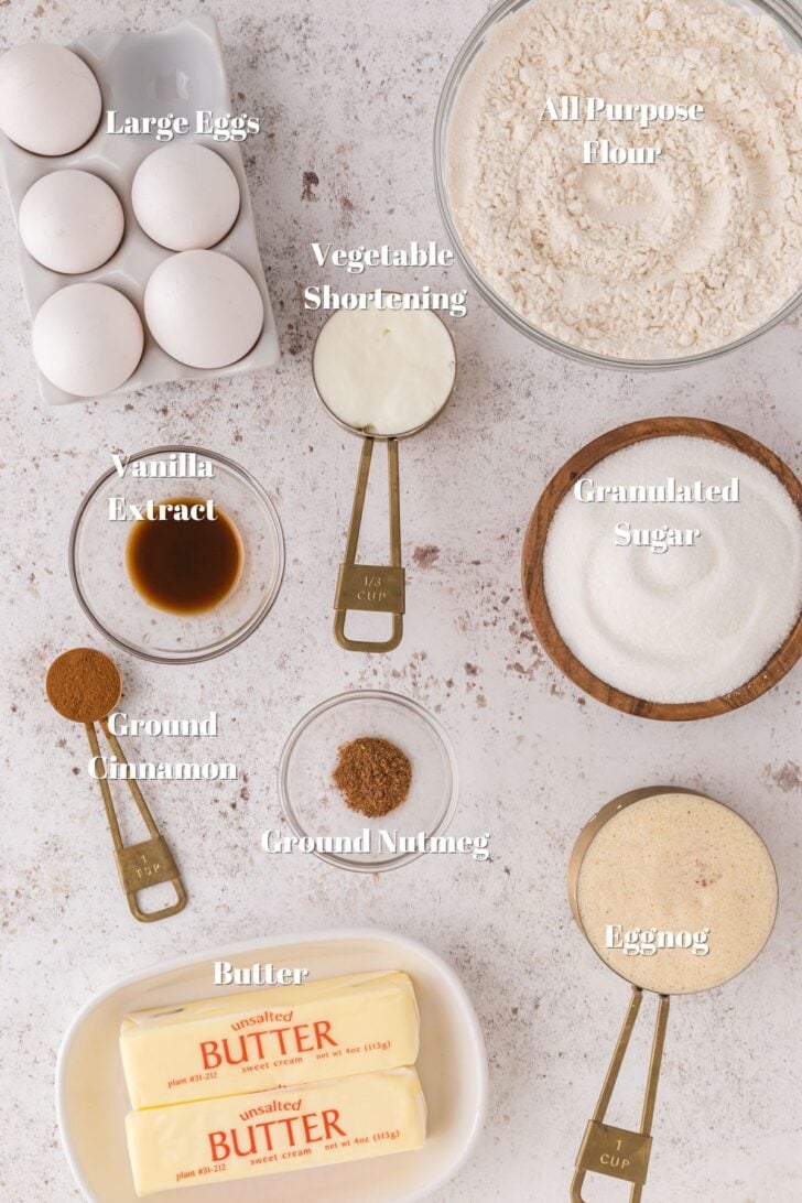 Large white eggs, all purpose flour in a glass bowl, vegetable shorting, vanilla extract in a glass bowl, granulated sugar in a wooden bowl, ground cinnamon, ground nutmeg, eggnog and sticks of butter on a white counter.