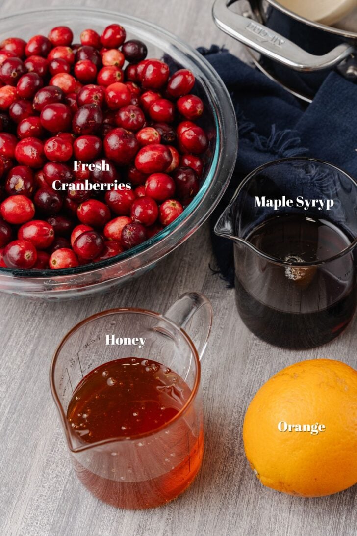 Fresh bowl of cranberries, glass measuring cup of maple syrup, glass measuring cup of honey and a whole orange on a wooden board.