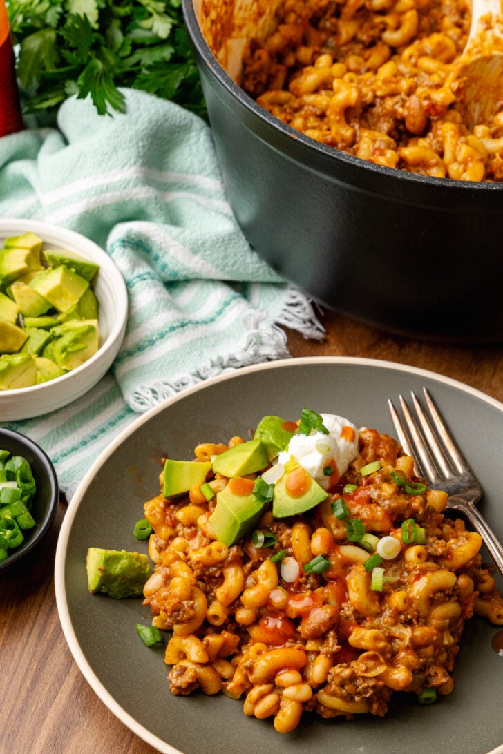Gray plate with chili mac topped with a dollap of sour cream, hot sauce, chopped green onion, and avocado served with a silver fork and a large pot with chili mac.