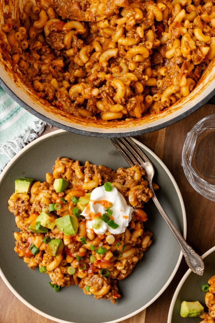 Gray plate with chili mac topped with a dollap of sour cream, hot sauce, chopped green onion, and avocado served with a silver fork and a large pot with chili mac.