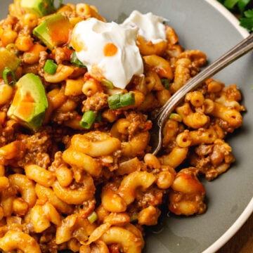 Gray plate with chili mac topped with a dollap of sour cream, chopped green onion, and avocado served with a silver fork.