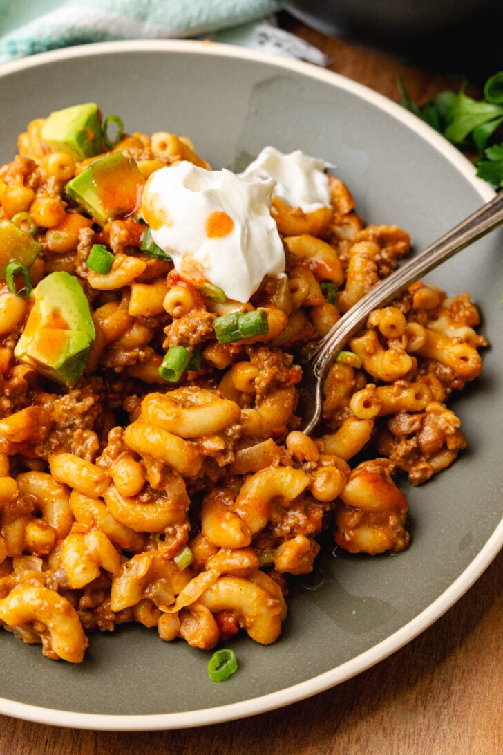 Gray plate with chili mac topped with a dollap of sour cream, chopped green onion, and avocado served with a silver fork.
