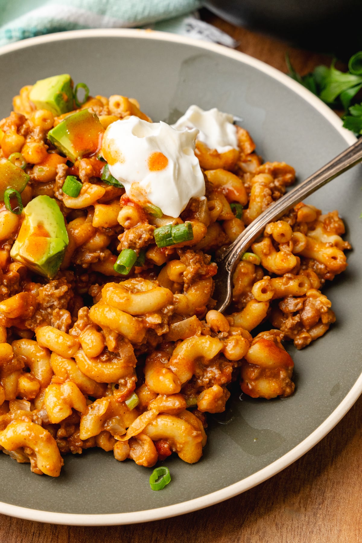 Gray plate with chili mac topped with a dollap of sour cream, chopped green onion, and avocado served with a silver fork.