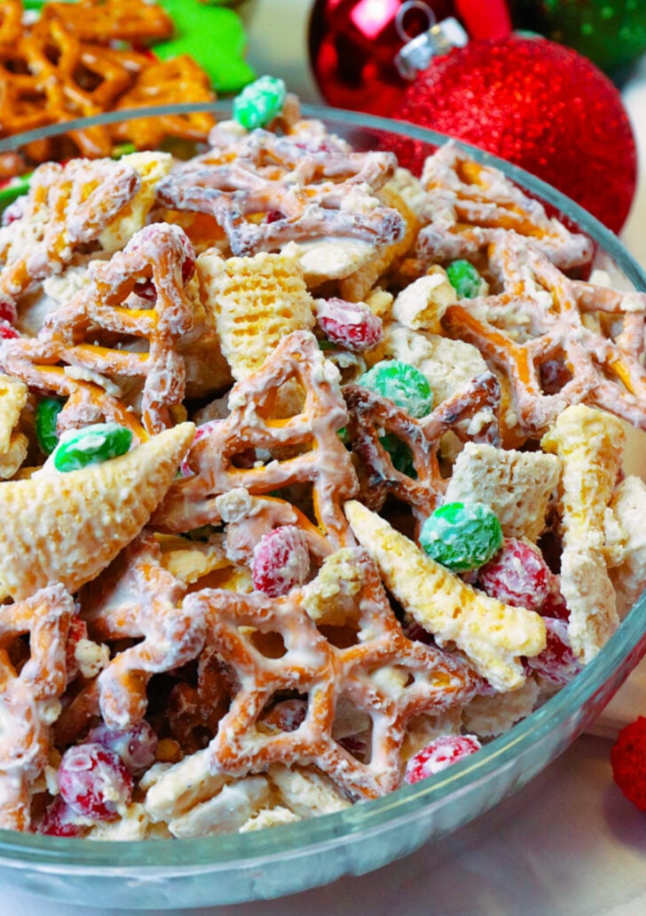 White vanila melted candy coating pretzels, rice cereal, bugles and M&M candy in a glass bowl.