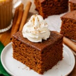 Gingerbread cake cut into a square topped with fresh whipped cream and sprinkled with cinnamon on a white plate with a silver fork and served with coffee.