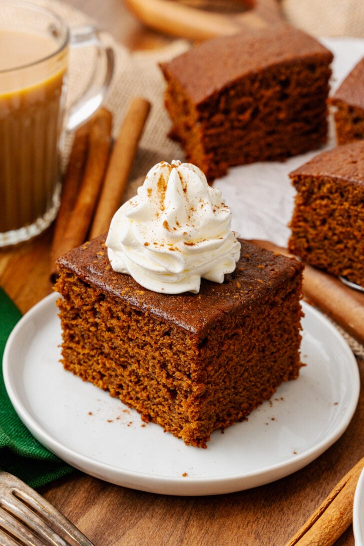 Gingerbread cake cut into a square topped with fresh whipped cream and sprinkled with cinnamon on a white plate with a silver fork and served with coffee.
