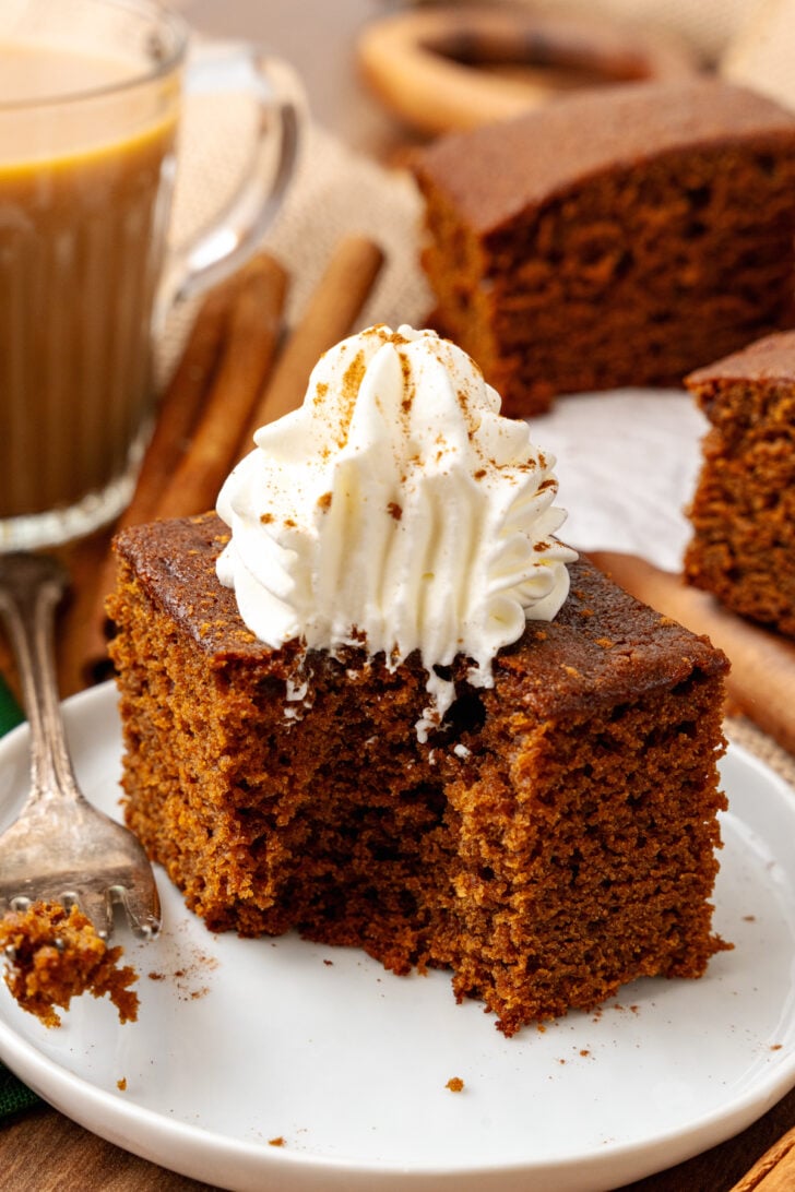 Gingerbread cake cut into a square topped with fresh whipped cream and sprinkled with cinnamon on a white plate served with a silver fork.