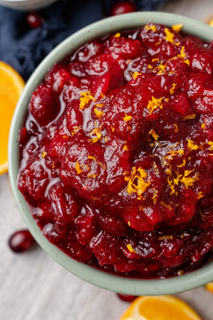 Small light green bowl of orange cranberry relish topped with fresh orange zest resting on a wooden board.