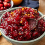 Small light green bowl of orange cranberry relish topped with fresh orange zest served with a silver spoon resting on a wooden board.