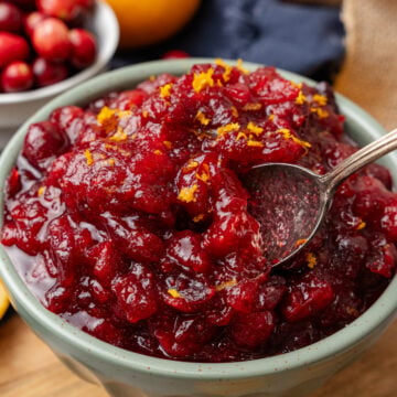 Small light green bowl of orange cranberry relish topped with fresh orange zest served with a silver spoon resting on a wooden board.