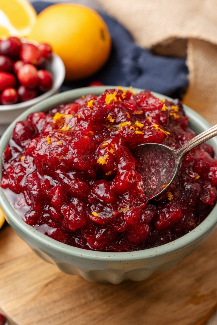 Small light green bowl of orange cranberry relish topped with fresh orange zest served with a silver spoon resting on a wooden board.