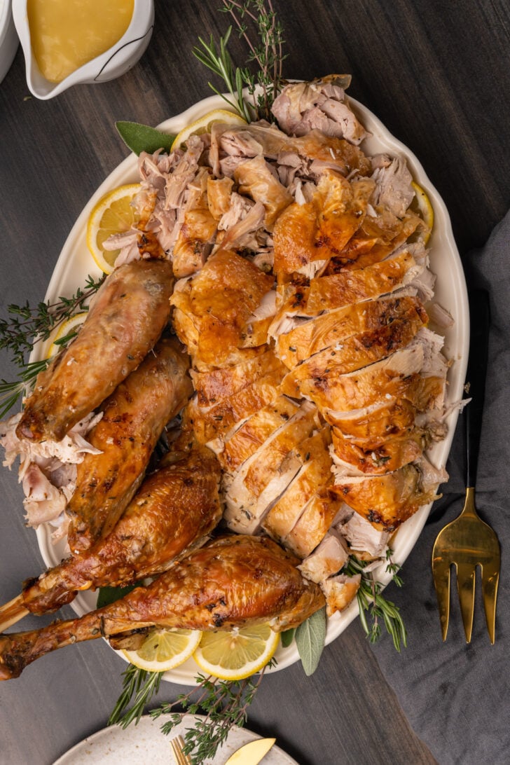 Carved roasted turkey breast, legs and wings garnished with fresh herbs placed on a white platter.