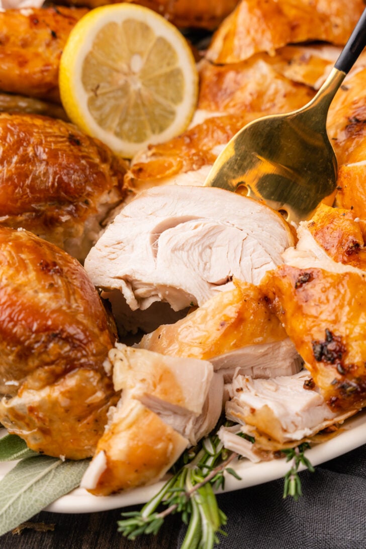 Roasted turkey slices on a white platter served with a gold fork garnished with fresh herbs and lemon slices.