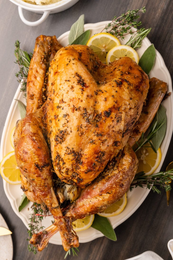 Whole roasted turkey on a white platter with fresh sage, thyme, and rosemary sprigs and lemon slices.