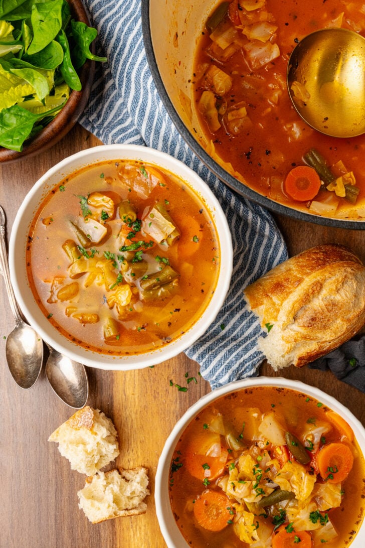 Two hite bowl will garden vegetable soup with carrots, green beans and cabbage topped with chopped parsley and a large bowl with soup all served with bread.