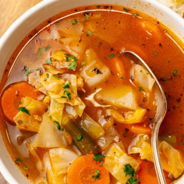 White bowl will garden vegetable soup with carrots, green beans and cabbage topped with chopped parsley and served with a silver spoon.