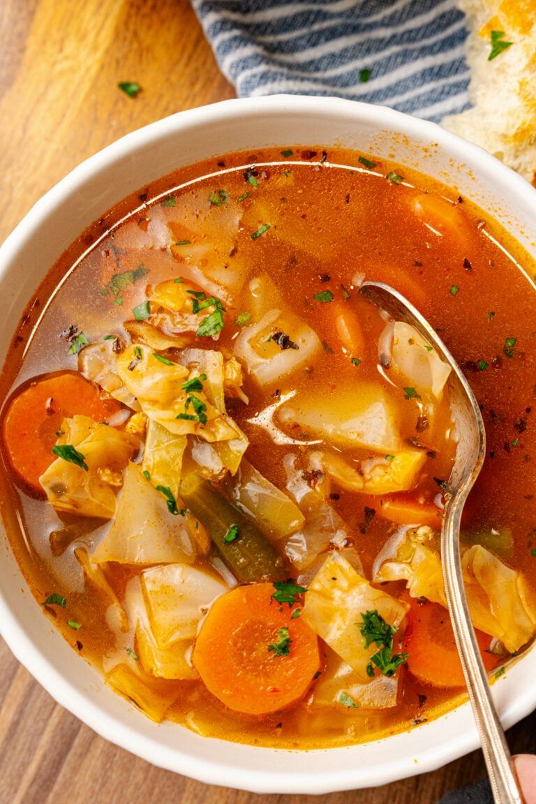 White bowl will garden vegetable soup with carrots, green beans and cabbage topped with chopped parsley and served with a silver spoon.