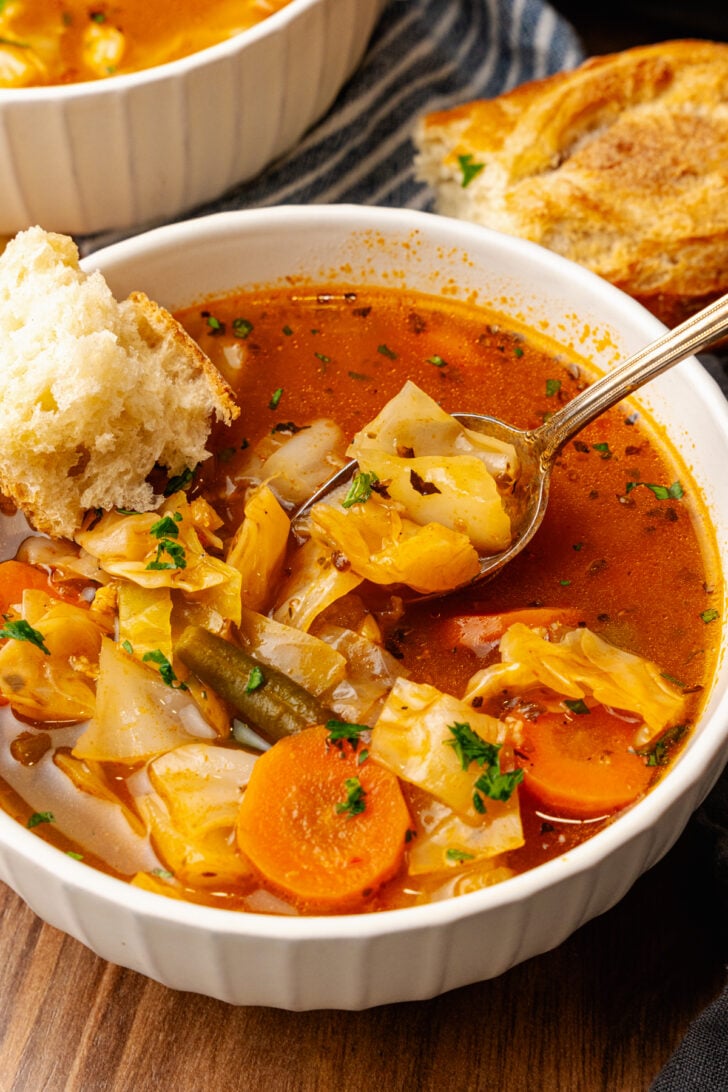 White bowl will garden vegetable soup with carrots, green beans and cabbage topped with chopped parsley and served with a silver spoon and a chunk of bread.