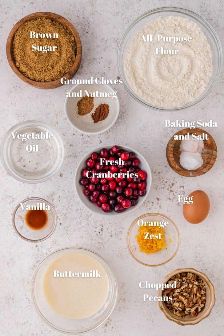 Brown sugar, all-purpose flour, ground nutmeg and cloves, vegetable oil, fresh cranberries, baking soda and salt, vanilla, buttermilk, orange zest, egg, and chopped pecans in bowls on a white counter.