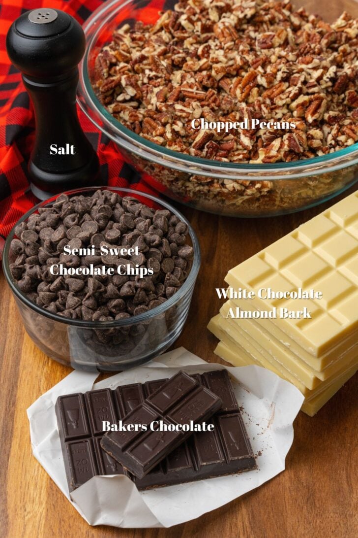Black Salt shaker, glass bowl with chopped pecans, glass bowl with semi sweet chocolate chips, white chocolate almond bark, and bakers chocolate on a wooden counter.