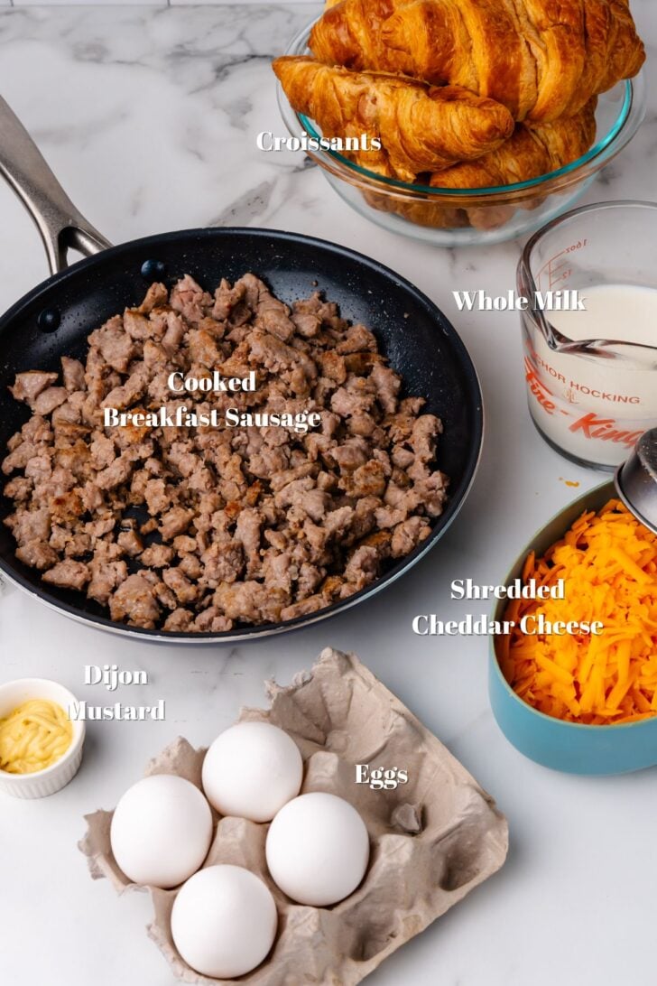 Croissants, cooked sausage, whole milk, shredded cheddar cheese, eggs and dijon mustard, ingredients on a white counter for breakfast casserols recipe.