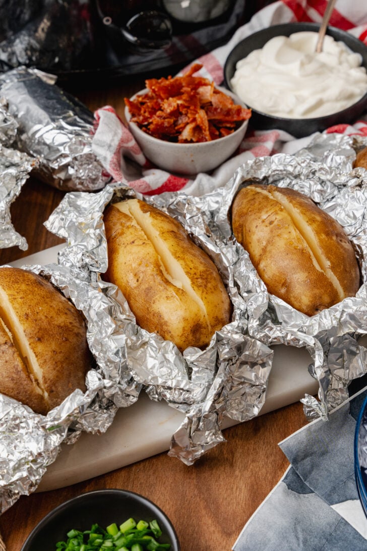 Baked potatoes split in half wrapped in aluminum foil ready to be stuffed with sour cream bacon cheese and chives.