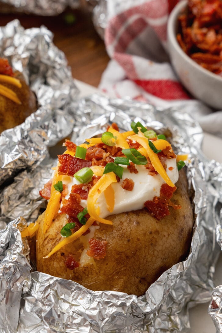 Baked potatoes topped with sour cream cheese bacon bits and chives on topo of a piece of aluminum foil.