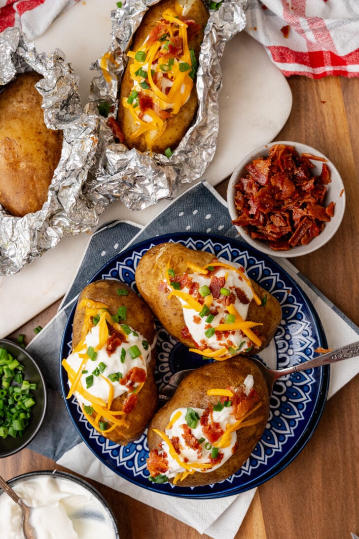 Baked potatoes topped with sour cream cheese bacon bits and chives ontop of a nice blue plate with all the fixing surrounding it.