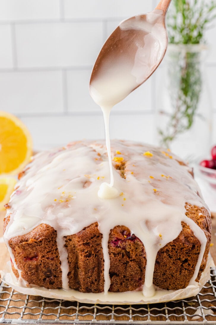 Metal spoon drizzling white glaze on cranberry orange quick bread resting on a metal rack.