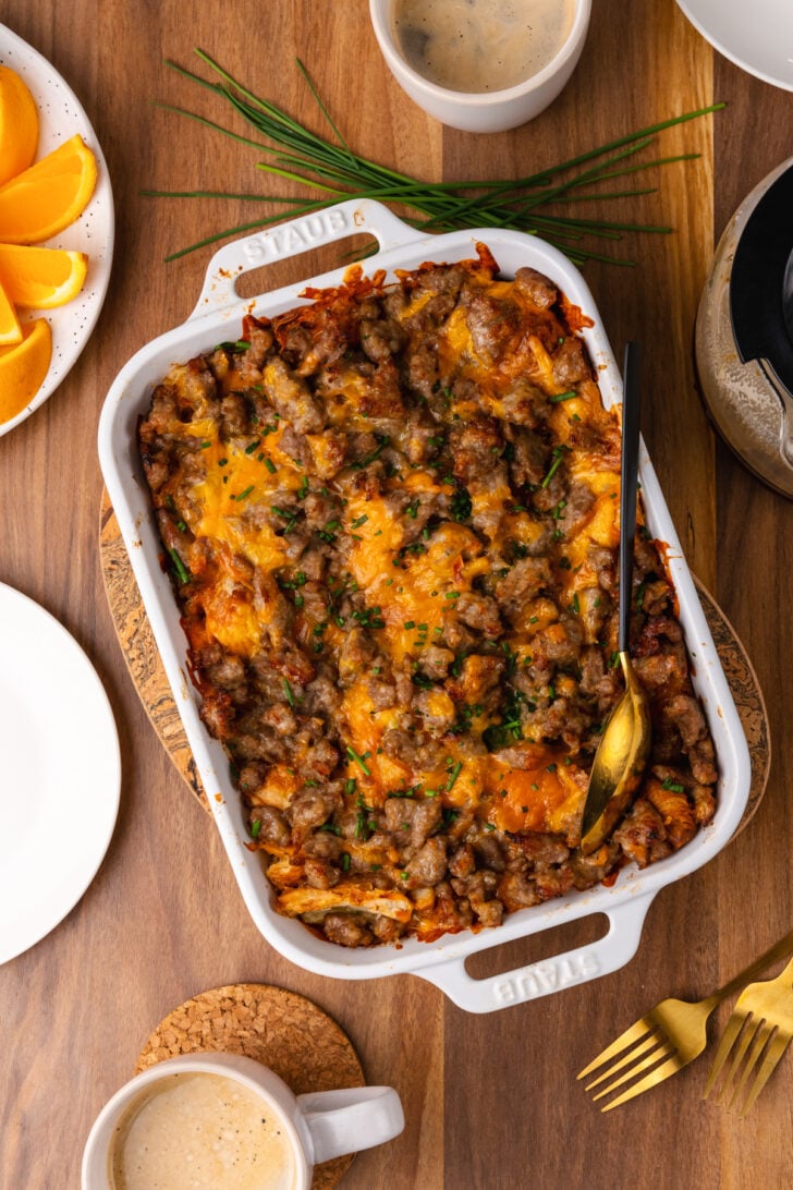 White cast iron baking dish with sausage and cheese croissant casserole topped with chives and served with a gold spoon, coffe and orange slices.
