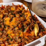 White cast iron baking dish with sausage and cheese croissant casserole topped with chives and served with a gold spoon.