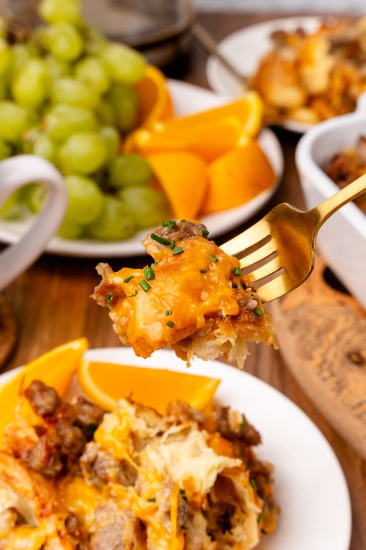 Gold fork with a bite of sausage and cheese croissant breakfast casserole taken from a white plate with casserole and orange slices.