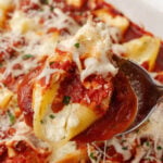 Spoon full of stuffed shells, stuffed with cheese parsley and marinara sauce inside a baking dish.