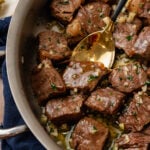 Metal saute pan with steak bites coated in butter, garlic and fresh parsley tossed with a gold spoon.