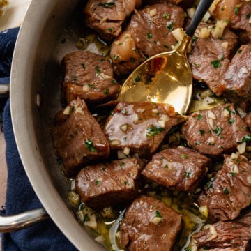 Metal saute pan with steak bites coated in butter, garlic and fresh parsley tossed with a gold spoon.