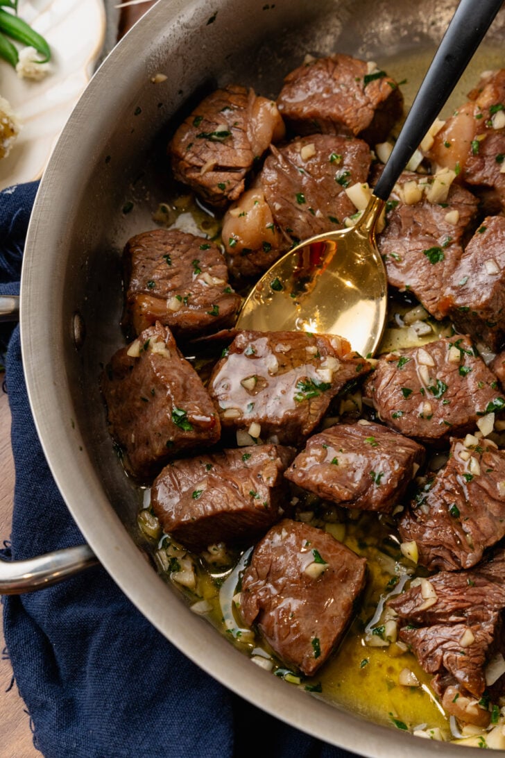 Metal saute pan with steak bites coated in butter, garlic and fresh parsley tossed with a gold spoon.