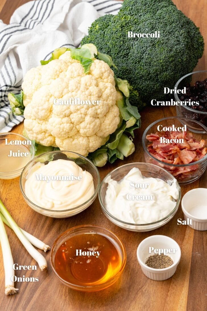 Broccoli, cauliflower, dried cranberries, cooked bacon, lemon juice, mayonnaise, sour cream, salt, pepper, green onion and honey in clear glass bowls on a wooden counter.