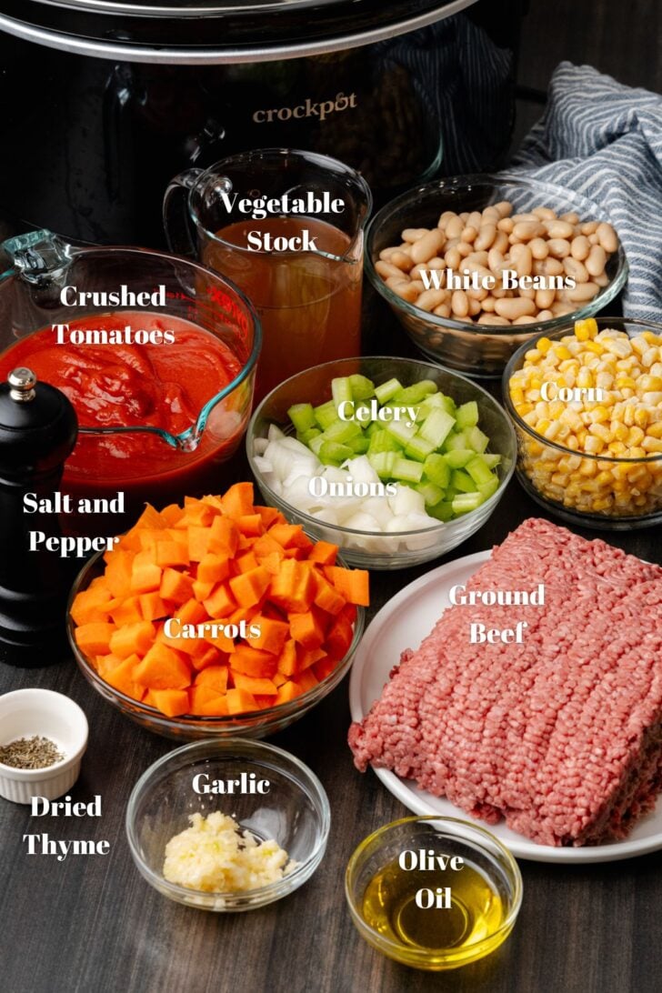 Crock pot, beans, stock, corn, ground beef, olive oil, garlic, thyme, carrots, onions, celery, crushed tomatoes, and vegetables in glass bowls for soup recipe.