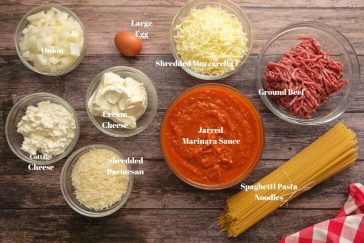 Ingredients of onion, egg, mozzarella, ground beef, pasta, marinara sauce, cream cheese, shredded sheese, and cottage cheese in glass bowls.