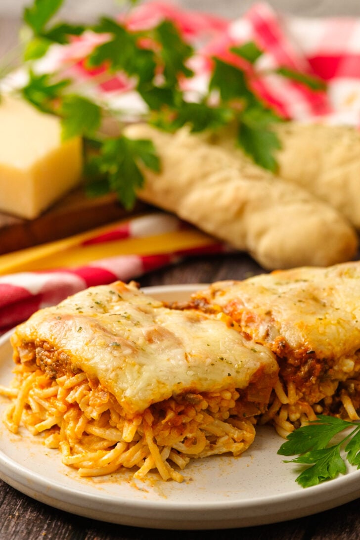 Baked spaghetti sliced on a white plate with parsley served with bread sticks.