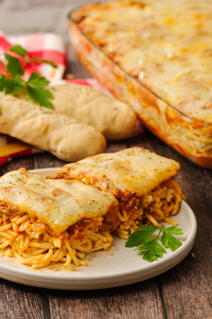 Baked spaghetti sliced on a white plate with parsley and a full casserole dish.
