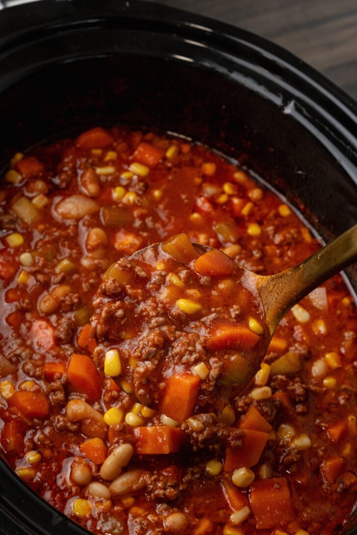 Black crock pot with beef and bean soup ladled with a brass spoon.