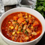 White bowl with ground beef, vegetables and bean soup on a wooden board with a silver spoon and fresh parsley.