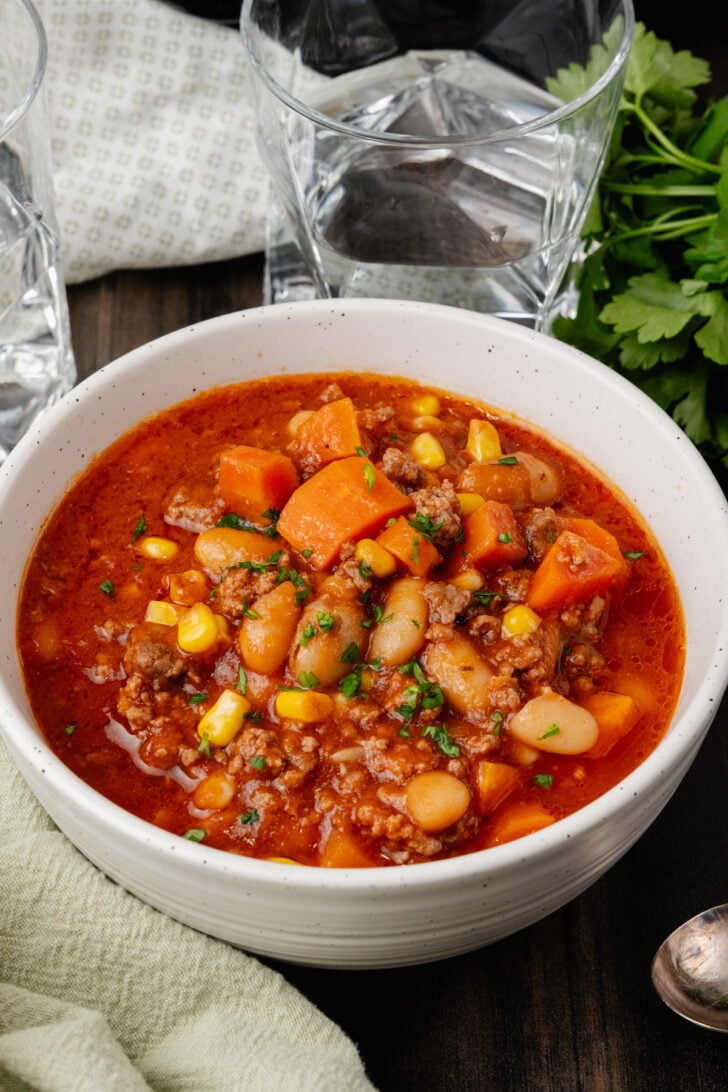White bowl with ground beef, vegetables and bean soup on a wooden board with a silver spoon and fresh parsley.