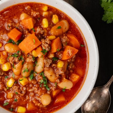 White bowl with ground beef, vegetables and bean soup on a wooden board with a silver spoon and fresh parsley.