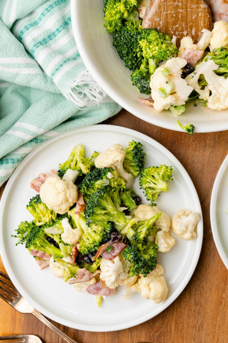 White bowl filled with broccoli cauliflower salad with cooked bacon and dried cranberries and a white plate with salad.