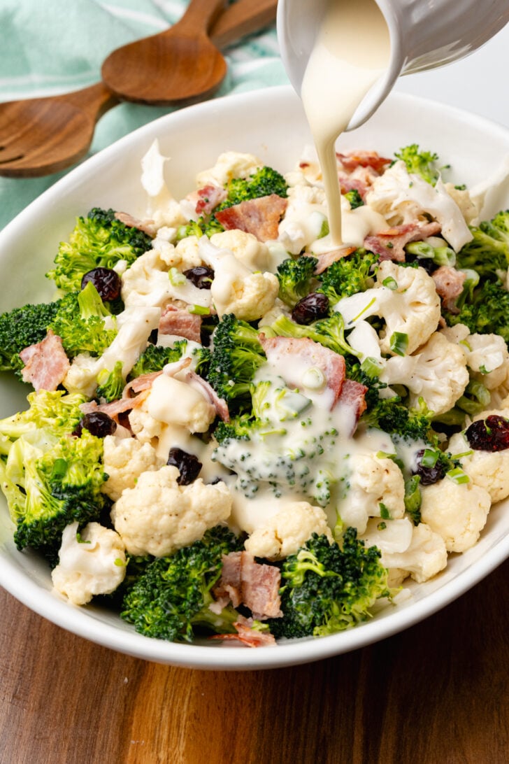 White bowl filled with broccoli cauliflower salad with cooked bacon and dried cranberries with creamy dressing poured over.