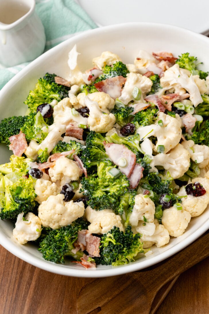 White bowl filled with broccoli cauliflower salad with cooked bacon and dried cranberries.