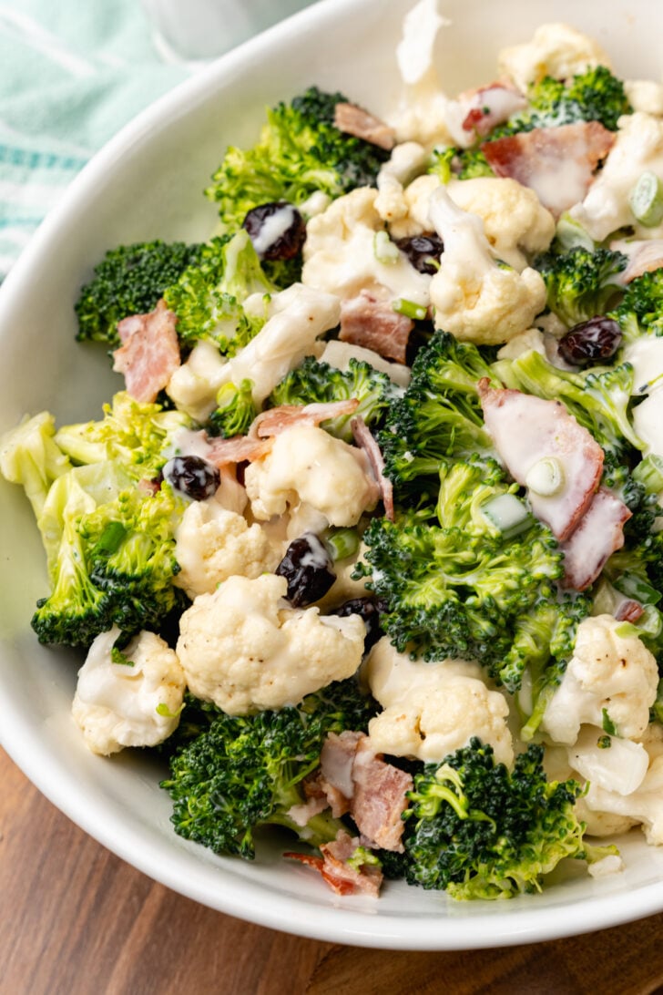 White bowl filled with broccoli cauliflower salad with cooked bacon and dried cranberries.